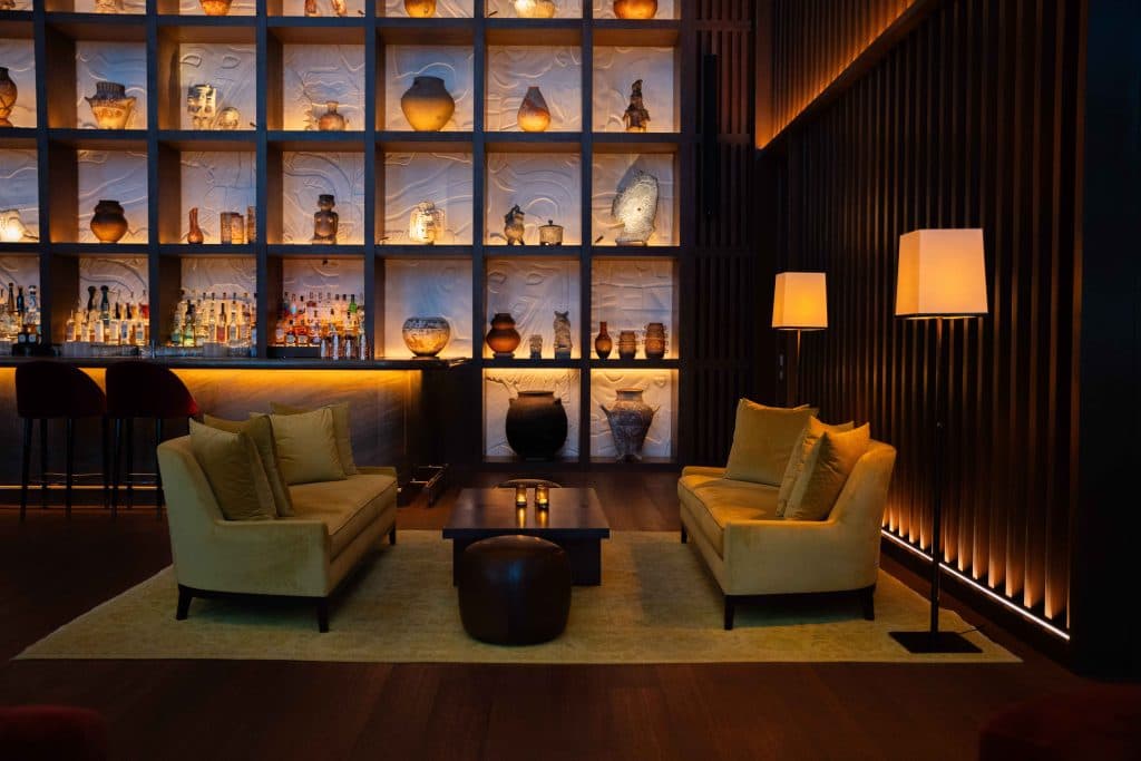 A hotel bar with lit shelving and velvet armchairs