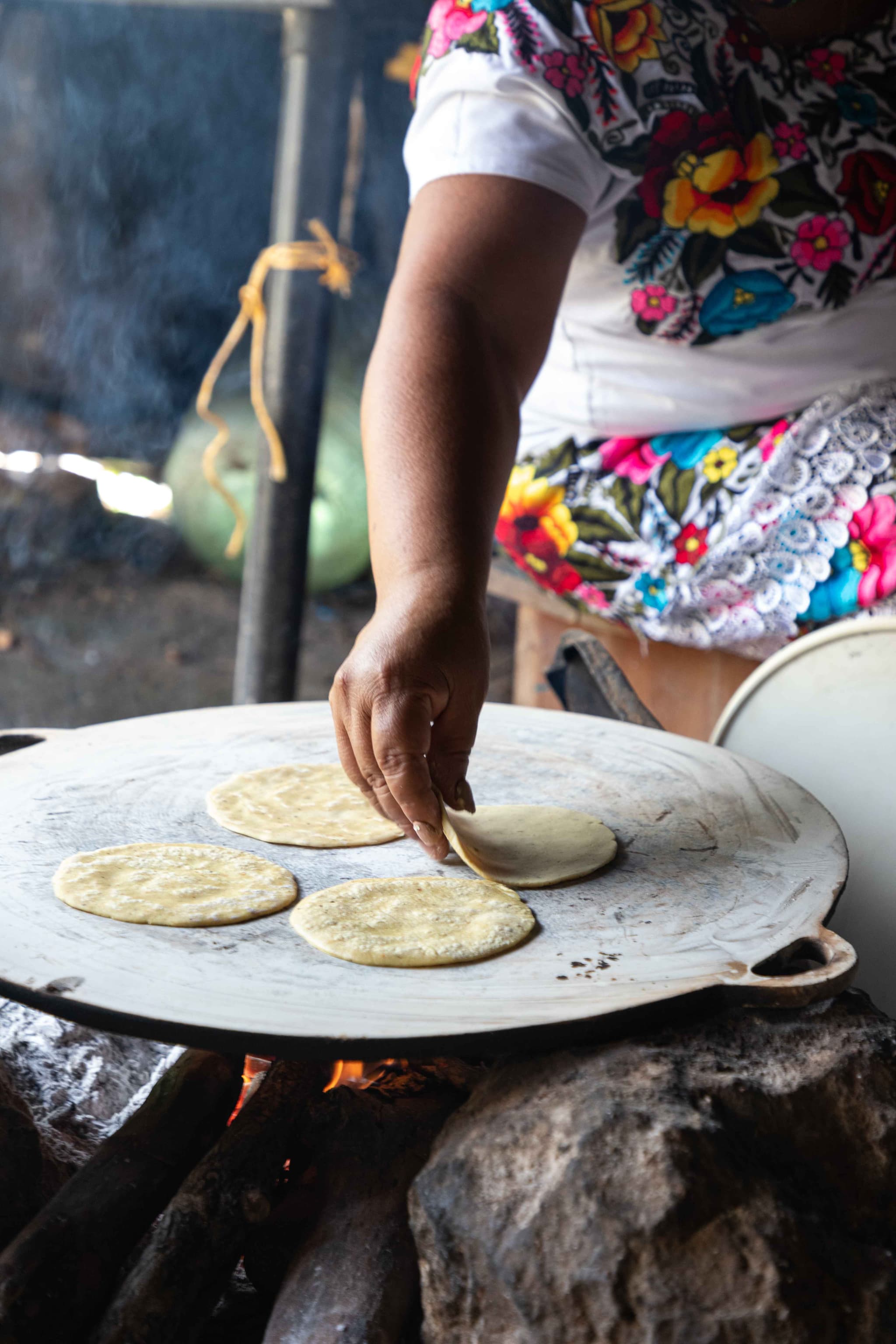 A cook in an embroidered huipil turning corn tortillas on a comal over a wood fire