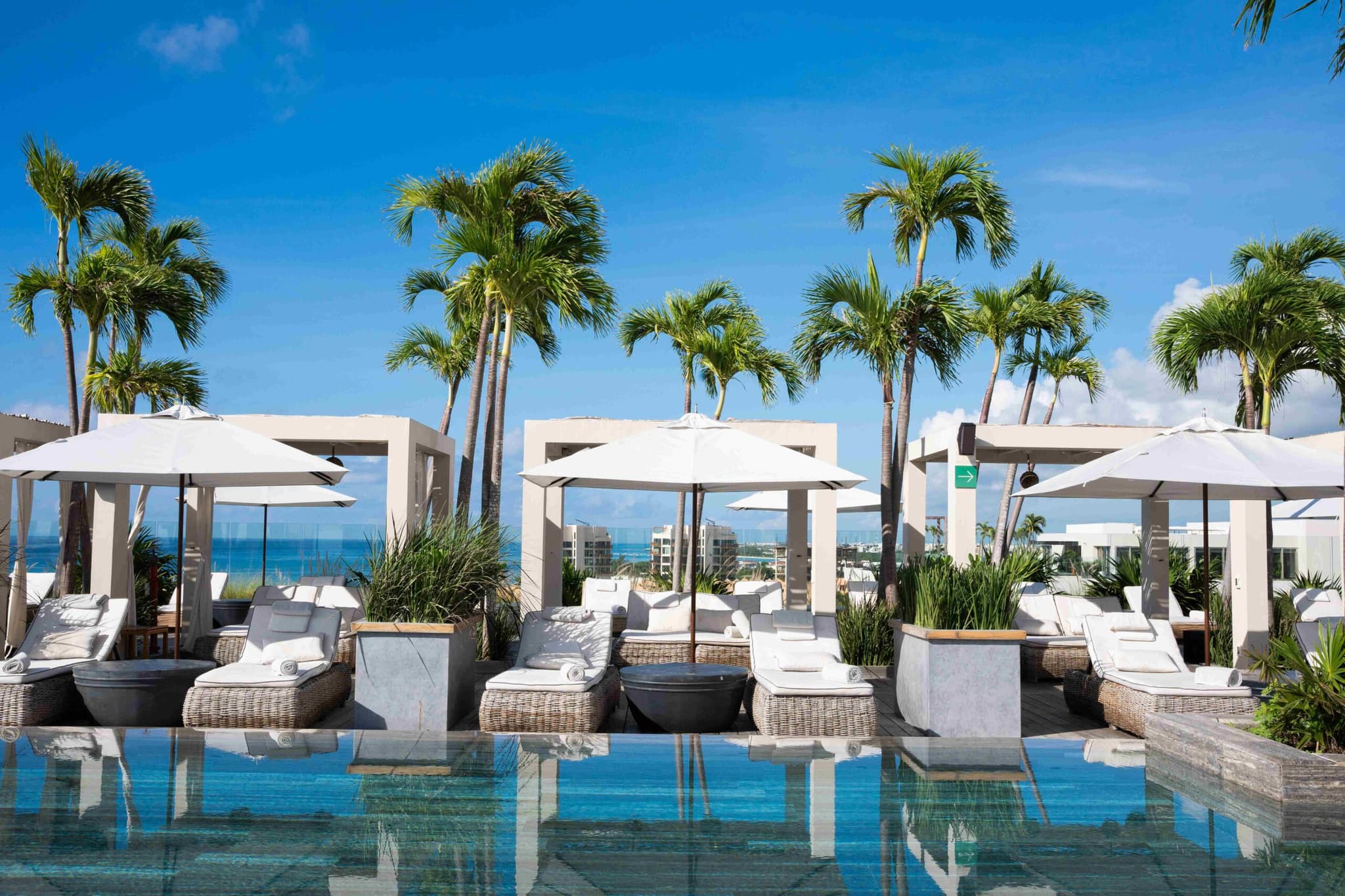 Loungers and white parasols at the pool edge, palms and the sea behind