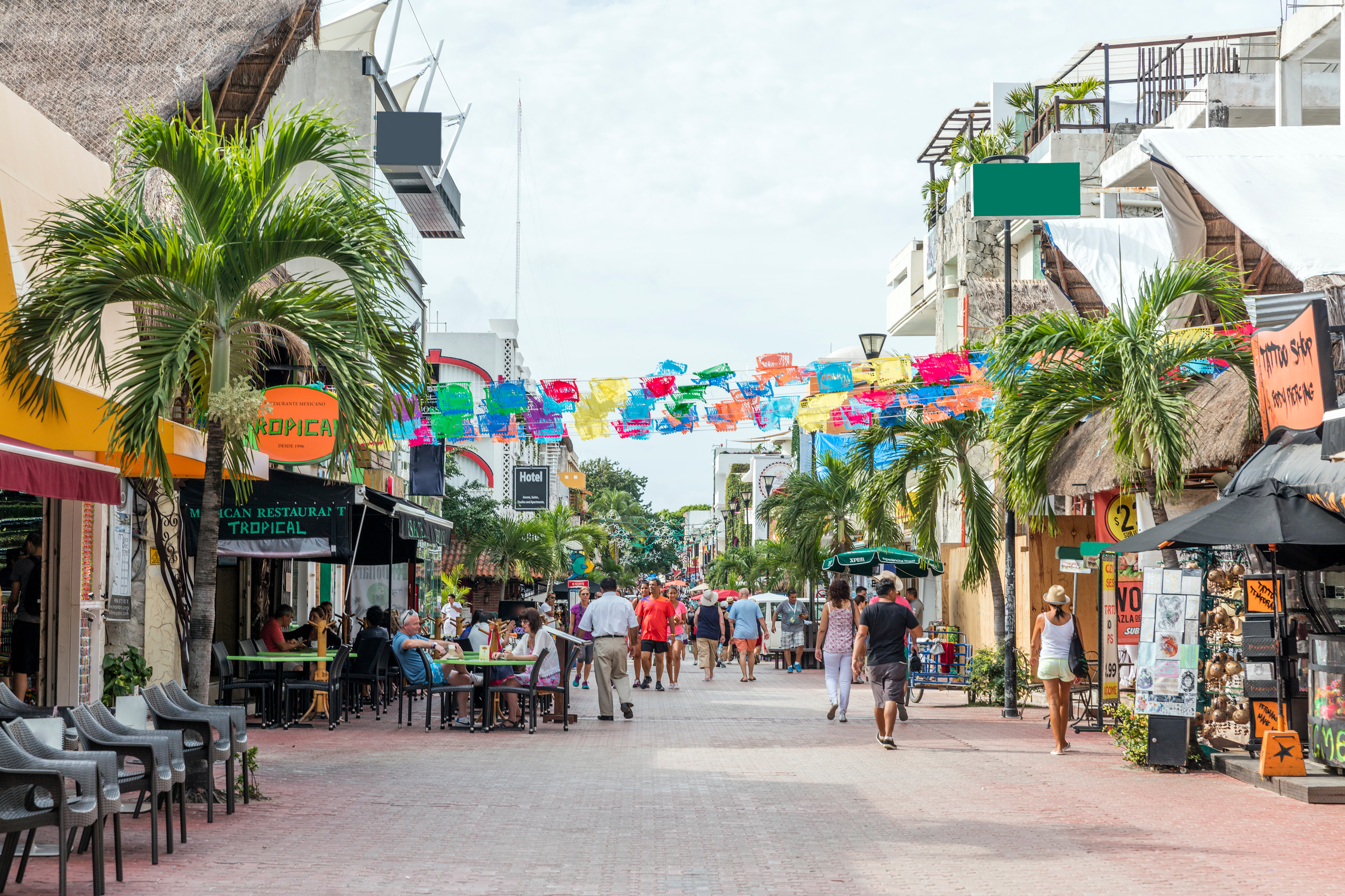 Fifth Avenue in Playa del Carmen by day, with papel picado banners between the palms, terraces and people strolling